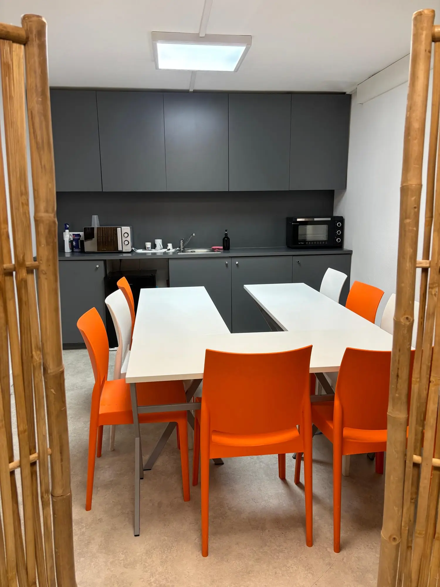 Espace cafétéria : grande table blanche, chaises orange, cuisine équipée derrière un claustra en bambou