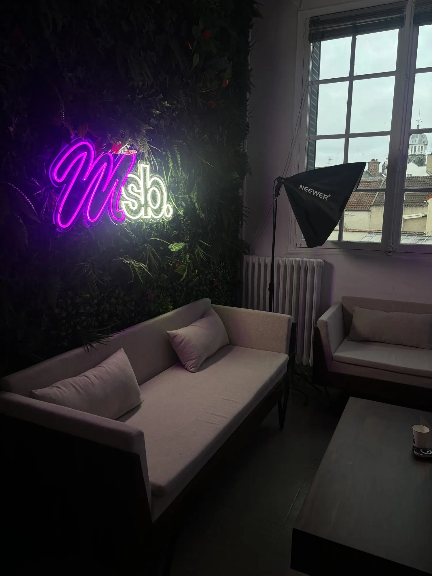 Studio d'enregistrement : néon rose au monogramme de l'école, mur végétal, éclairage de tournage et canapé